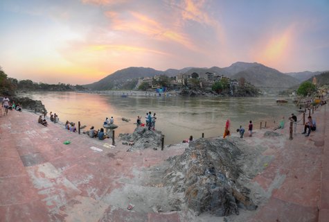 Wide panoramic view of the Ganges River in Rishikesh at sunset with people sitting on stone ghats, buildings on hillsides, and mountains in the background under a pink and orange sky