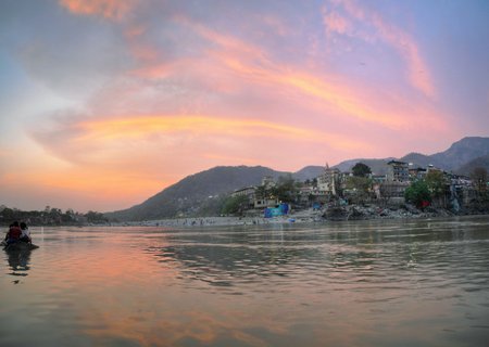 Wide view of Ganges River at sunset with pink and orange sky, buildings along the shore, mountains in background, and people sitting on rocks in the water