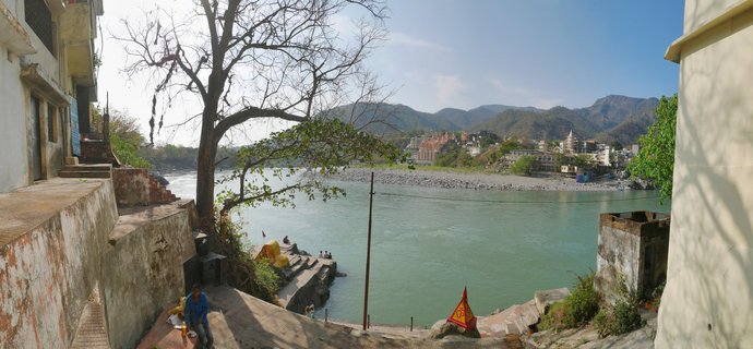 Wide view of the Ganges River in Rishikesh showing stone ghats, a large tree, riverside buildings, temples across the water, and mountains in the background under blue sky