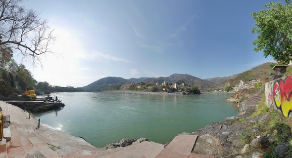 Wide view of the Ganges River in Rishikesh with stone steps leading to greenish-blue water, buildings and ashrams on both banks, and mountains in the background under a clear sky