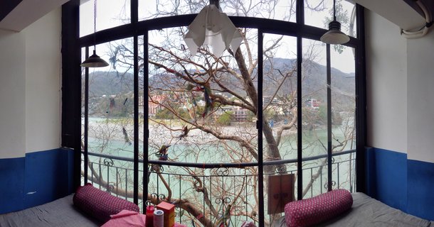 Room interior with large arched black-framed window overlooking Ganges River, bare tree with colorful prayer flags, turquoise water, buildings and hills visible across the river, two cushioned daybeds with red bolster pillows in foreground