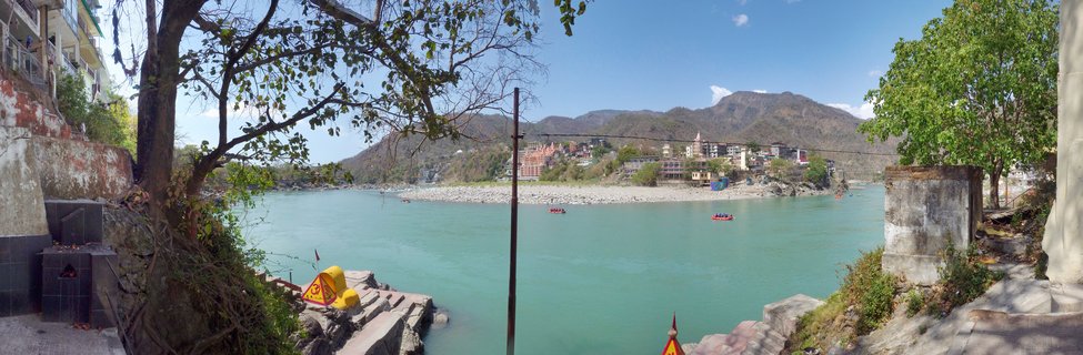 Panoramic view of the Ganges River in Rishikesh showing turquoise water with red rafting boats, riverside buildings and temples, stone steps leading to water, trees framing the scene, and mountains in the background under a clear blue sky