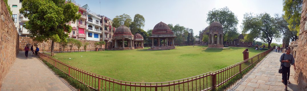Three domed Mughal pavilions with red sandstone columns on a green lawn, surrounded by stone walkways with visitors, trees, and modern buildings in the background at Hauz Khas Complex, Delhi