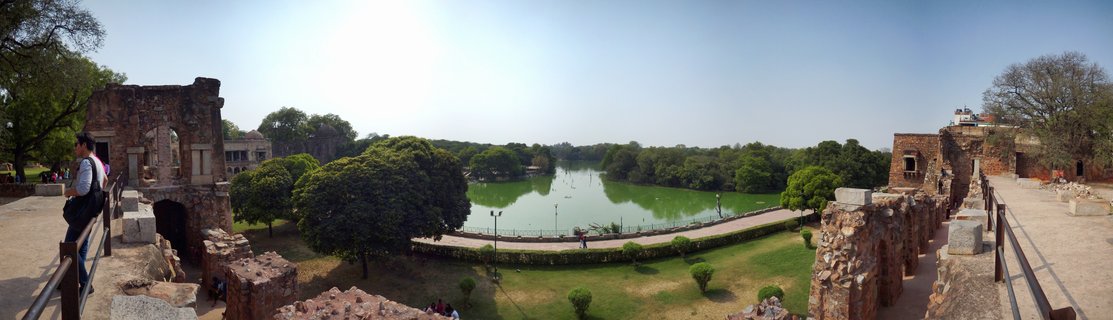 Panoramic view of ancient red sandstone fortress ruins on both sides framing a green lake with trees, pathways, and clear blue sky in Delhi, India