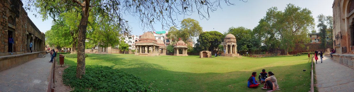 Historical domed stone pavilions and chhatris on a green lawn at an archaeological site in Delhi with visitors walking and sitting nearby