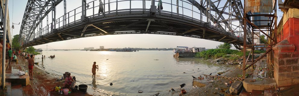 Steel cantilever bridge arching over wide river with people bathing at steps along the water's edge during sunset
