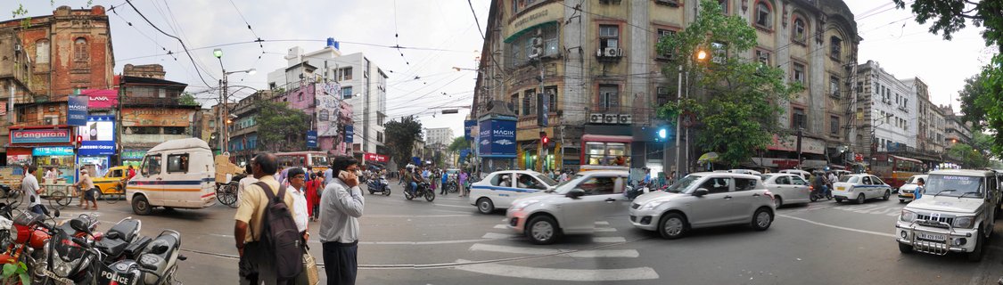 Wide panoramic street scene showing a busy intersection in Kolkata with multiple vehicles including cars, motorcycles, taxis, pedestrians crossing, colonial buildings, and commercial signage