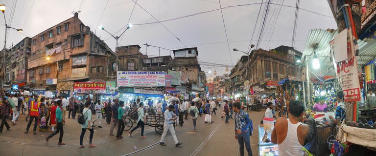 Wide street scene in Kolkata showing numerous pedestrians walking among old buildings with shop signs, tram tracks visible on the road surface, overhead electrical wires crisscrossing above