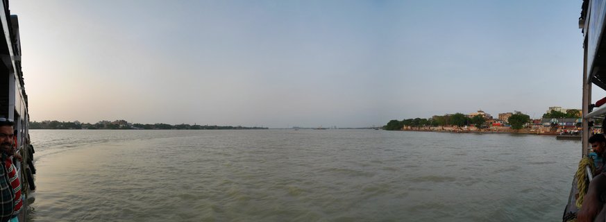 Wide river view from a ferry boat showing the Hooghly River in Kolkata with boat structures visible on the left and right edges, passengers on deck, riverbanks with buildings and trees in the distance, and golden afternoon sky above the water