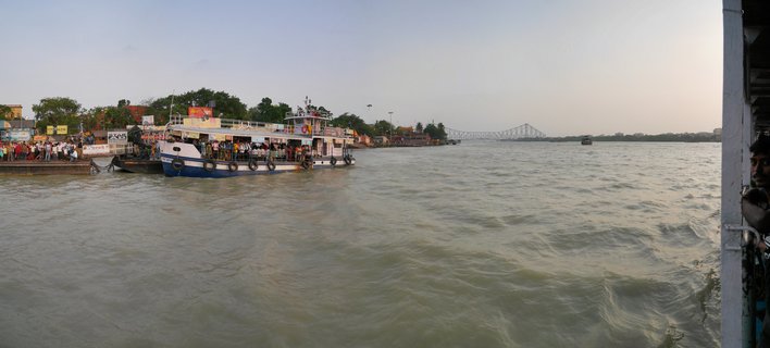 A crowded passenger ferry docked at a river ghat on the Hooghly River with people waiting on the platform and the Howrah Bridge visible in the background under a hazy late afternoon sky