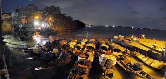 Night photograph of multiple wooden cargo boats docked at a river ghat with people, crates, and barrels under street lighting in Kolkata