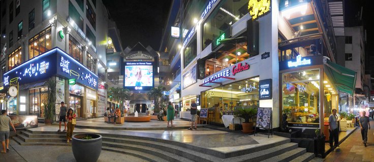 Night photograph of an outdoor shopping plaza in Bangkok showing illuminated cafe storefronts with neon signs in Korean and English, a large LED screen displaying welcome message, and people walking on paved courtyard with steps and planters