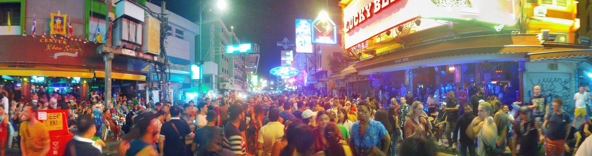 Crowded street scene at night showing hundreds of people walking on Khao San Road in Bangkok with neon signs, bars, restaurants, and Thai flags visible on buildings
