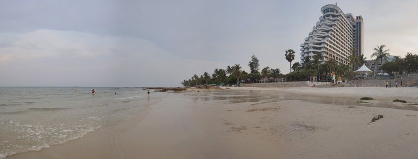 Wide beach scene showing people swimming in ocean water on the left, sandy beach in foreground, and a large white curved hotel building with palm trees on the right side under overcast sky