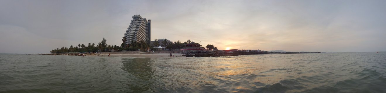 Panoramic beach scene at sunset showing Centara Grand Beach Resort with curved architecture, palm trees lining the sandy shore, people on the beach, and calm ocean water with golden sunset reflections under a warm hazy sky in Hua Hin, Thailand