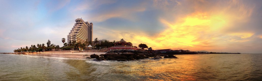 Panoramic coastal scene showing a large beachfront hotel resort, palm trees, sandy beach, rocky outcrops in shallow water, and golden sunset sky with dramatic clouds over the ocean in Hua Hin, Thailand