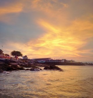 Sunset over coastal water in Thailand with golden sky, stilt buildings on pier, and rocky shoreline in foreground