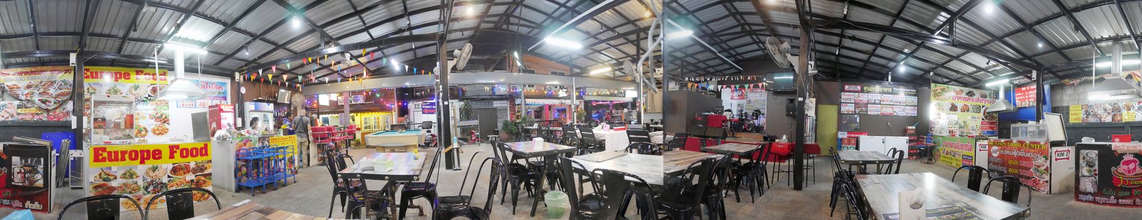 Interior view of a covered food court in Thailand showing multiple food vendor stalls with colorful signs, empty tables and chairs, and fluorescent lighting under a metal roof