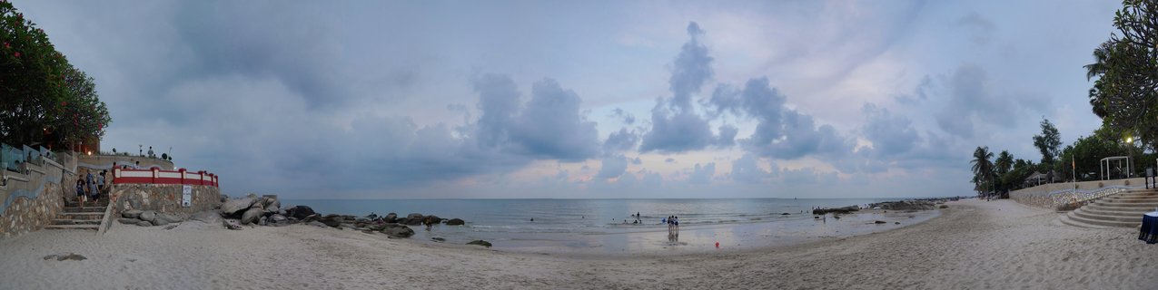 Wide beach panorama showing sand, calm ocean water, people walking and swimming, dramatic cloudy sky at dusk, stone structures with stairs on both sides, trees including palms