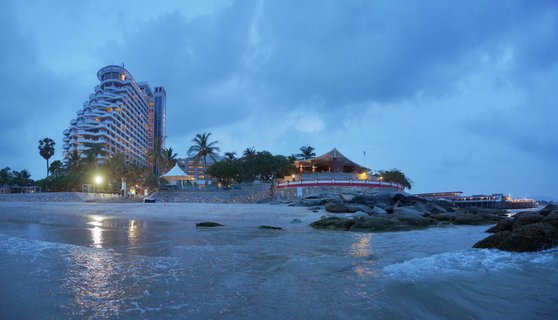 Beach resort at twilight with illuminated hotel building, palm trees, rocky shoreline, ocean waves, and pavilion structure under cloudy blue sky in Hua Hin, Thailand