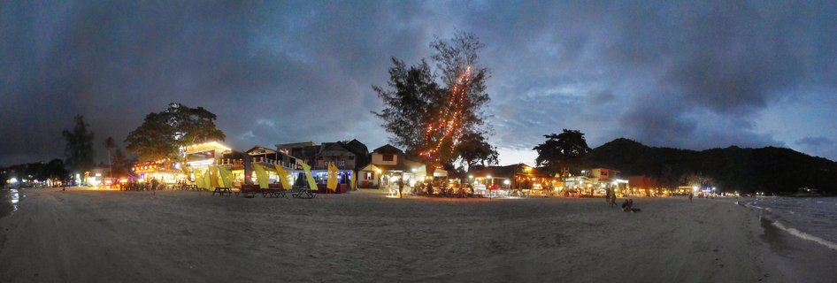 Panoramic view of illuminated beach bars and restaurants along a sandy beach at twilight with decorated trees, yellow flags, and people scattered on the sand under a cloudy blue evening sky