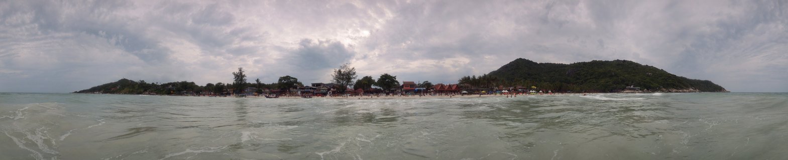 Panoramic beach scene showing a tropical bay in Thailand with resorts, palm trees, green hills, cloudy sky, and ocean waves in the foreground