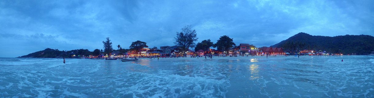 Wide beach panorama at twilight showing ocean waves in foreground, people on sand, illuminated beachfront restaurants with warm lights, long-tail boats, trees, and hills under cloudy blue evening sky in Koh Phangan, Thailand