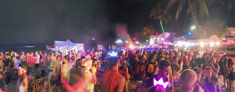 Large crowd of people at a nighttime beach party with colorful neon lights, DJ stages, palm trees, and glow accessories on Haad Rin Beach in Thailand