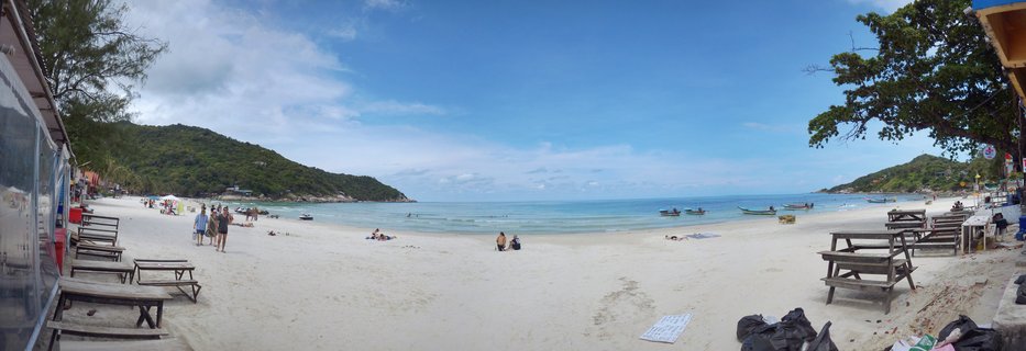 Panoramic view of a white sand beach in Thailand with turquoise ocean water, traditional longtail boats, scattered beachgoers, wooden tables, and green forested hills on both sides of the bay under a blue sky with clouds.