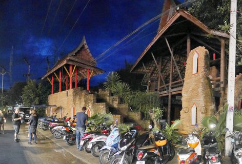 Night scene showing traditional Thai buildings with pointed roofs, red columns, and brick walls with multiple colorful motor scooters parked in front and tourists standing on wet pavement under a deep blue evening sky