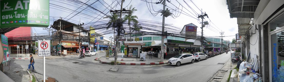 Panoramic street scene showing shops, palm trees, parked white cars, motorcycles, traffic signs with Thai script, and dense overhead electrical wires in Koh Samui, Thailand