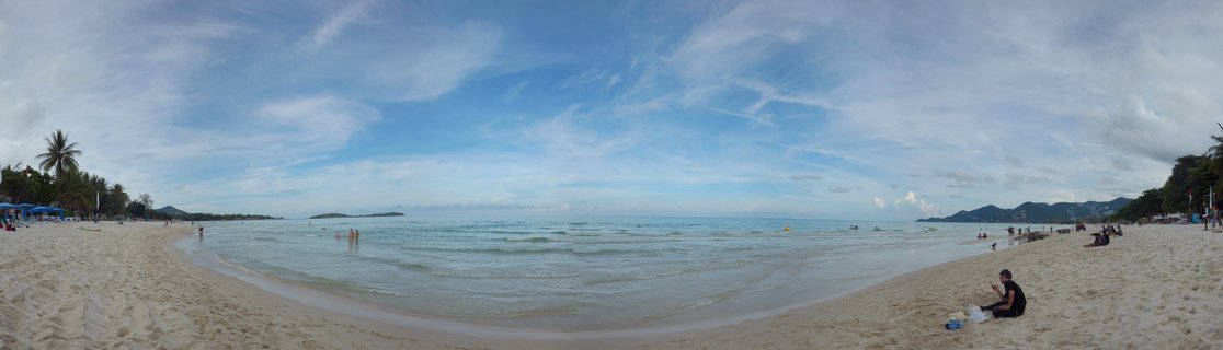 Wide beach panorama showing white sand, blue ocean water, palm trees on the left, people on beach and in water, distant mountains and islands on horizon under cloudy blue sky