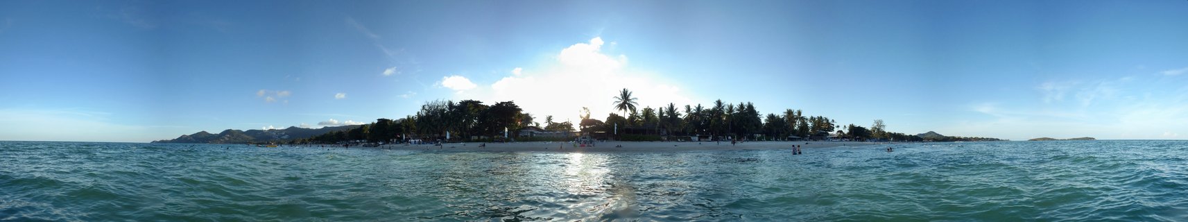 Panoramic photograph of a tropical beach in Koh Samui, Thailand showing turquoise ocean water in the foreground, white sand beach with palm trees and people in the middle ground, and distant mountains under a blue sky with scattered clouds