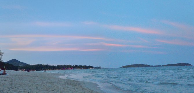 Panoramic beach scene at twilight showing white sand, ocean waves, pink clouds in blue sky, distant islands on horizon, and illuminated shoreline with trees and buildings along the coast in Koh Samui, Thailand