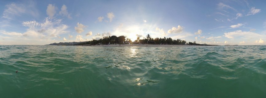 Panoramic view of turquoise ocean water with gentle waves, white sand beach with palm trees and buildings in the distance, blue sky with scattered clouds, and sun reflecting on the water surface at sunset