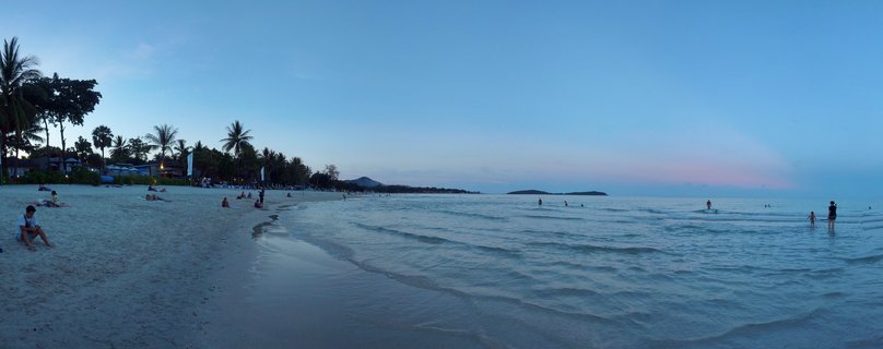 Wide beach scene at dusk in Thailand showing palm trees on the left, people on sand and in shallow water, calm ocean extending to distant islands under blue and pink sky
