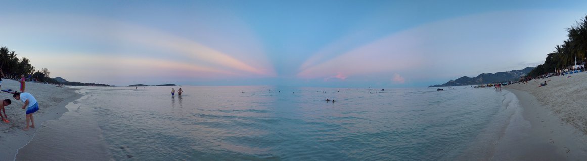 Panoramic view of a beach in Koh Samui, Thailand at sunset with crepuscular rays in a pink and blue sky, people swimming in calm ocean water, white sand beach with palm trees on both sides, and distant mountains on the horizon