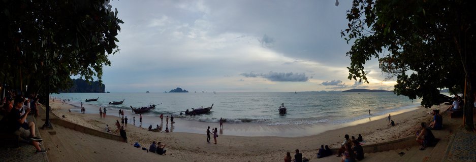 Wide beach scene showing tourists on sand, multiple long-tail boats in calm water, distant limestone islands, and overcast sky with trees framing both sides