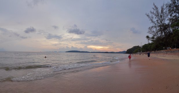 Wide beach scene at sunset showing people walking on wet sand, gentle waves, distant islands on horizon, trees along right side, cloudy sky with warm glow