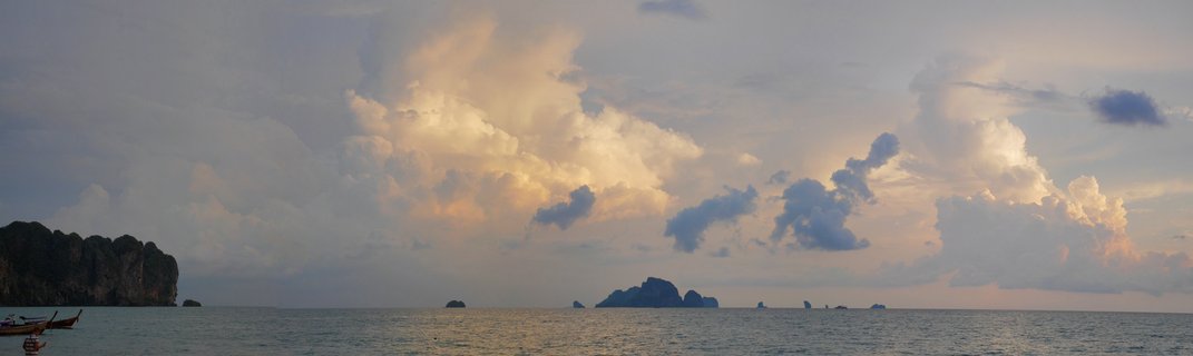 Panoramic seascape at sunset showing dramatic clouds over calm ocean water with limestone rock formations and traditional Thai long-tail boats at Ao Nang, Thailand