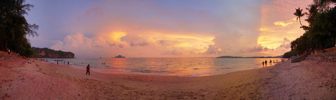 Wide panoramic beach scene at sunset showing golden and pink sky over ocean water, sandy beach with people walking, traditional longtail boat, limestone cliff formations on the left, and palm trees on the right side
