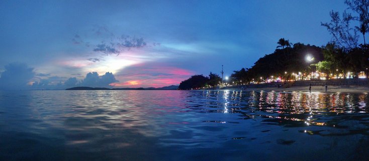 Panoramic beach scene at twilight showing pink and purple sunset sky over calm ocean water with reflections, beachfront lights and trees along the right shoreline, and silhouetted figures on the sand