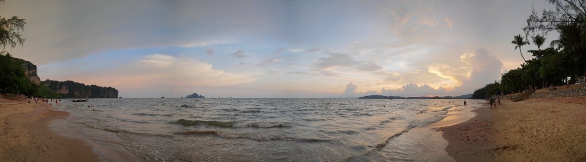 Wide beach scene at sunset showing Ao Nang Bay in Thailand with limestone cliffs on the left, tropical trees lining the shore, calm ocean waters, distant islands, and people walking on the sandy beach under a colorful evening sky