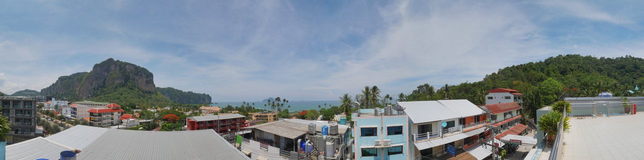 Panoramic aerial view of Ao Nang coastal town in Thailand showing buildings, palm trees, limestone mountain cliffs, and ocean under blue sky with clouds