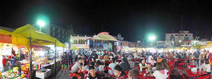 Crowded outdoor night market in Thailand with people dining at red tables, yellow food stall tents, and a stage structure under bright artificial lighting