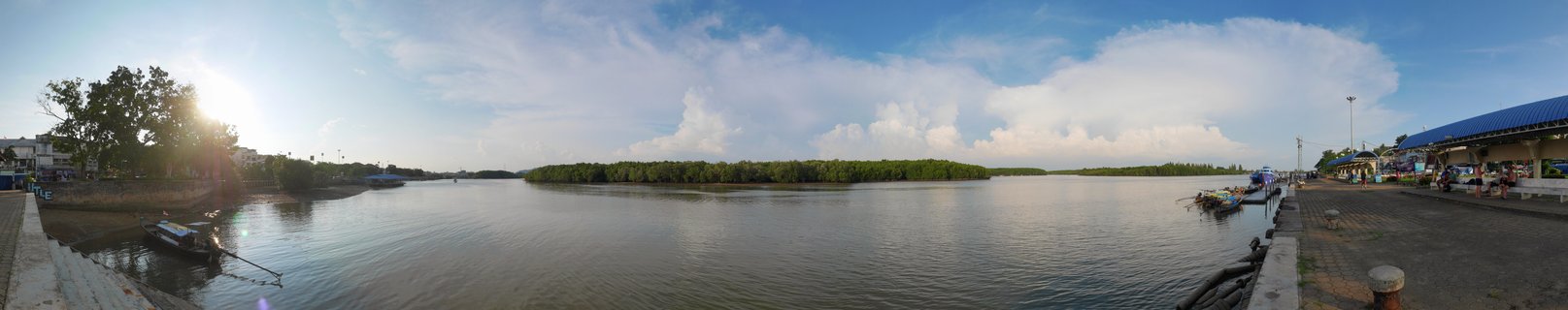 Panoramic waterfront scene showing a wide river estuary with calm brown water, traditional Thai long-tail boats docked at a concrete pier on the right, dense green mangrove forest on an island in the center distance, buildings and trees along the left bank, bright sun in the upper left sky with lens flare, blue sky with white clouds
