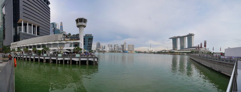 Wide panoramic photograph of Singapore's Marina Bay showing Marina Bay Sands hotel, modern skyscrapers, waterfront piers, and calm bay waters under cloudy sky