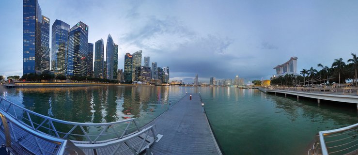 Panoramic view of Singapore Marina Bay at dusk showing illuminated skyscrapers, Marina Bay Sands hotel, wooden pier with person, and building reflections in water