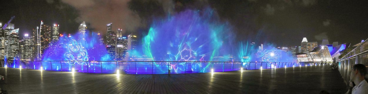 Night panorama of Marina Bay Spectra light and water show with colorful blue and purple water projections, Singapore CBD skyline illuminated in background, wooden boardwalk foreground with viewing spectators