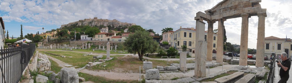 Panoramic view of the Gate of Athena Archegetis ancient ruins in Athens with the Acropolis hill visible in the background, showing stone columns, archaeological site grounds, olive trees, traditional buildings, and tourists exploring the historic location under a partly cloudy sky